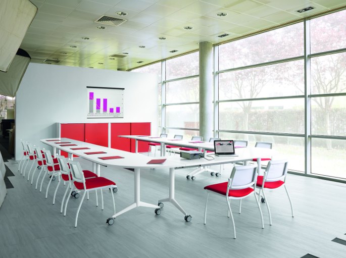 Salle de formation lumineuse avec tables alignées face à un écran et sièges rouges
